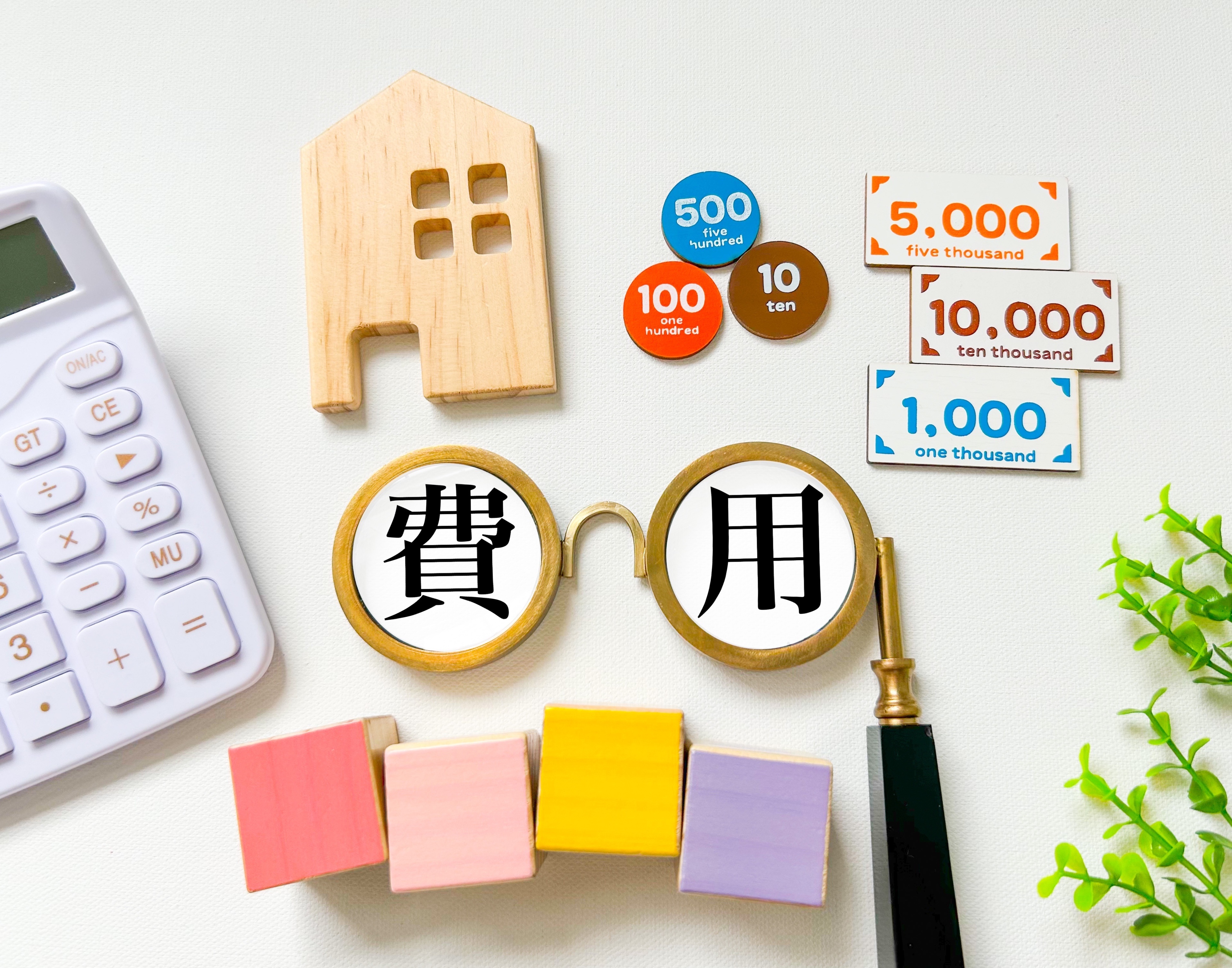 丸眼鏡のレンズ内に「費」「用」と書かれたオブジェを中心に、おもちゃのお金、木製の家、電卓などが並ぶ住宅費用のイメージ