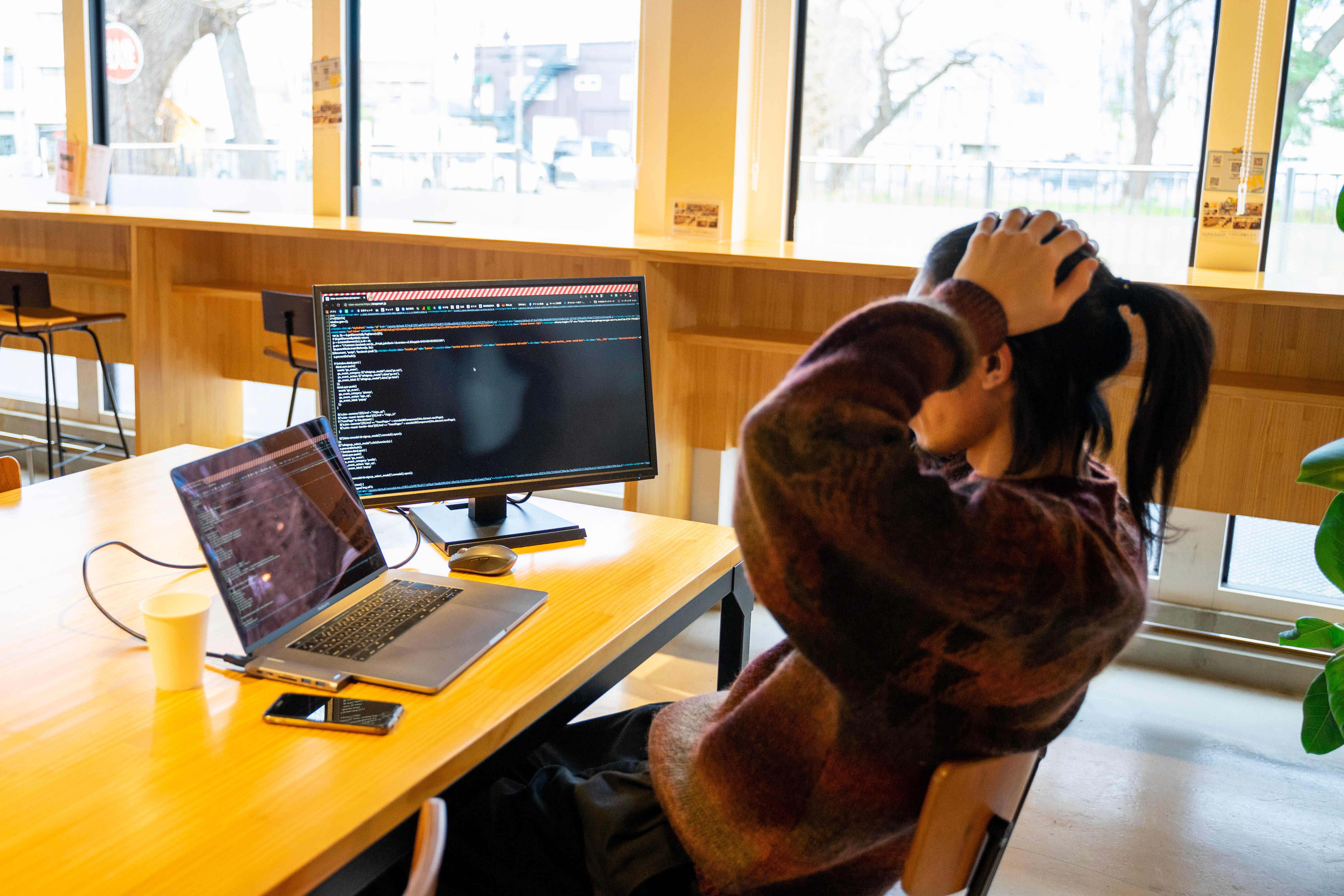 プログラミングコードが表示されたモニターとパソコンを前に、両手で頭を抱えて作業に行き詰まっている開発者の後ろ姿