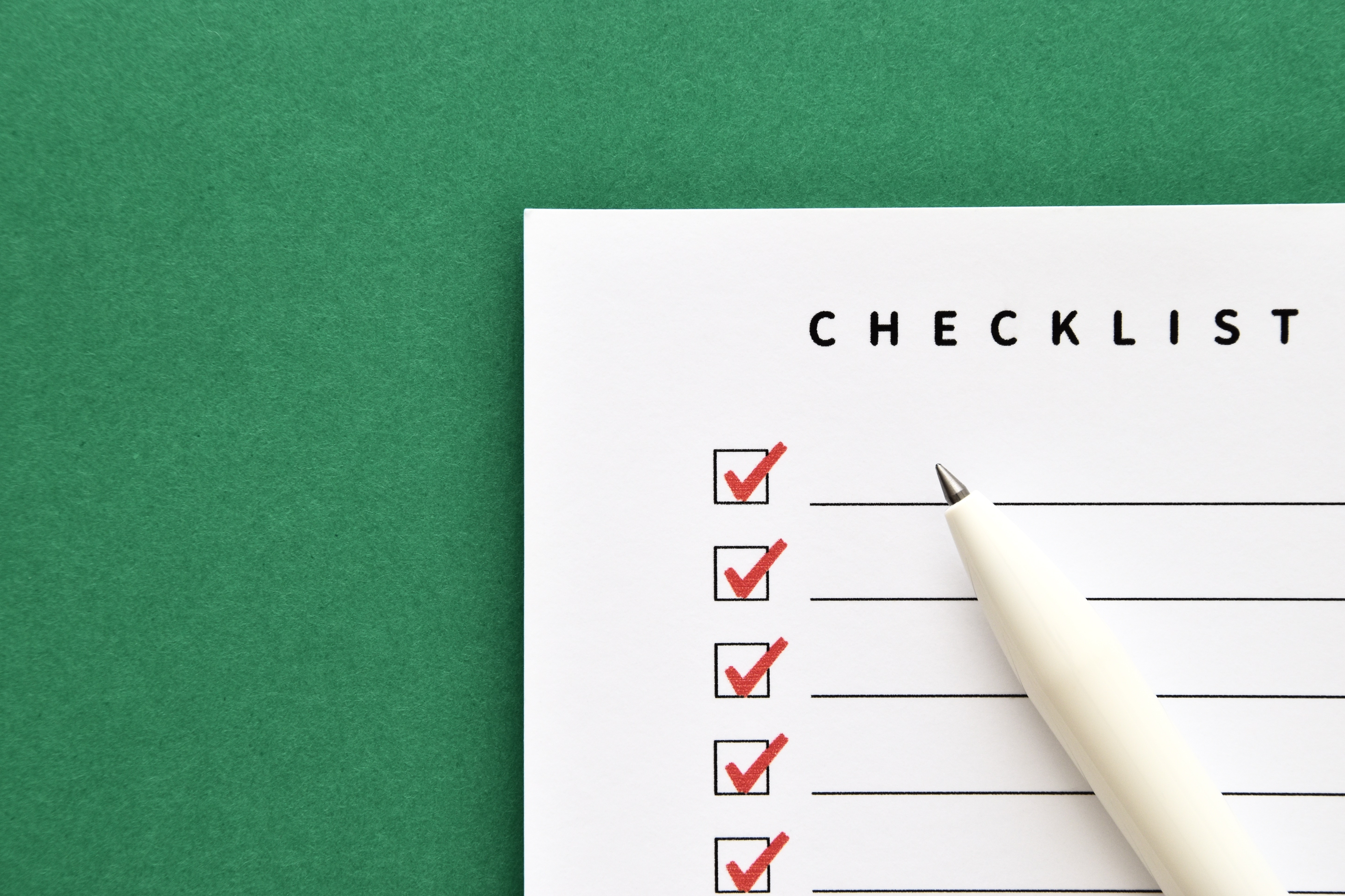 緑色の背景の上に置かれた「CHECKLIST」の白い用紙。すべてのチェックボックスに赤いチェックマークが入っており、白いペンが添えられている