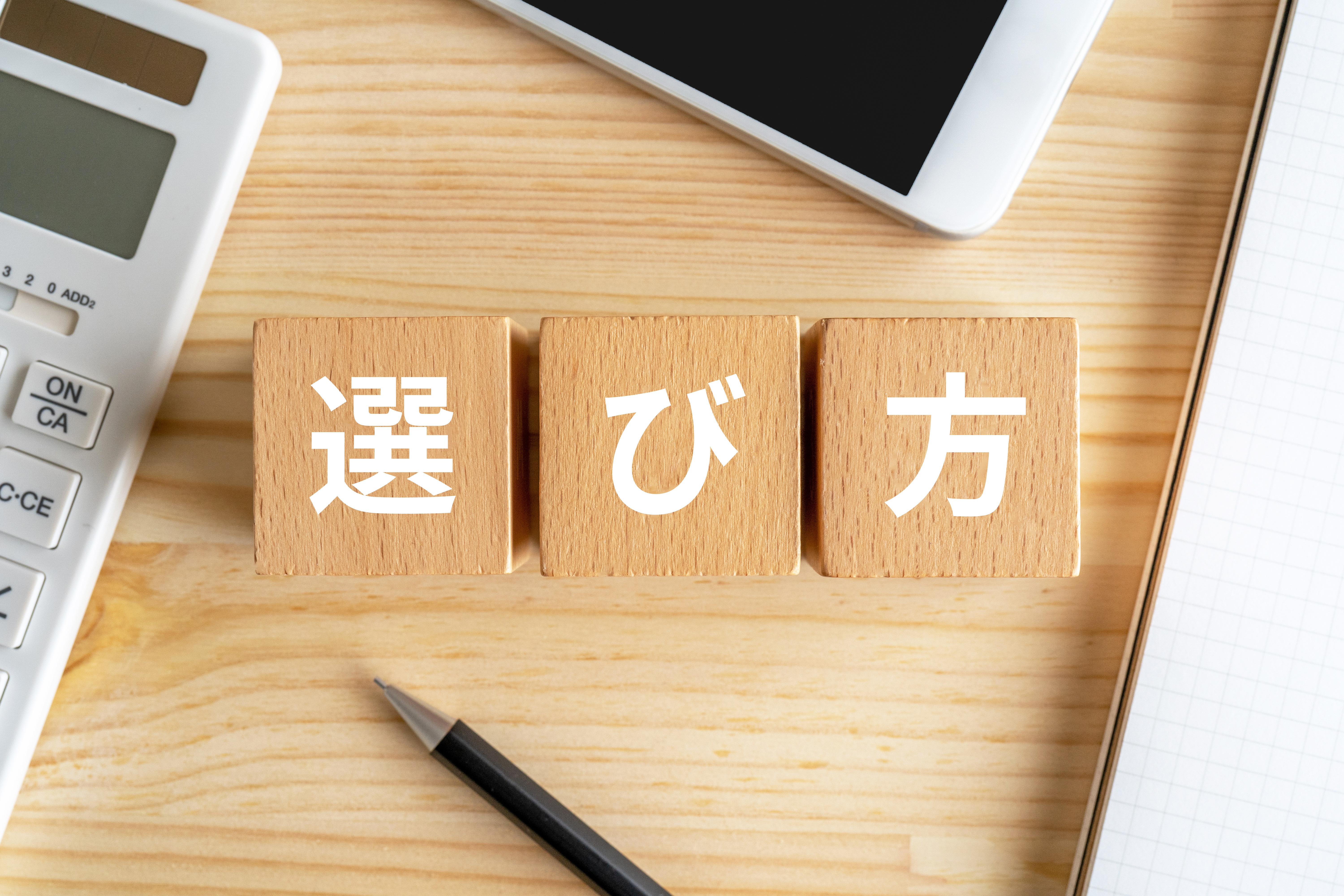 「選び方」の文字が書かれた木製ブロック、周囲に配置された電卓、スマートフォン、ノート、ペンを上から撮影した俯瞰イメージ